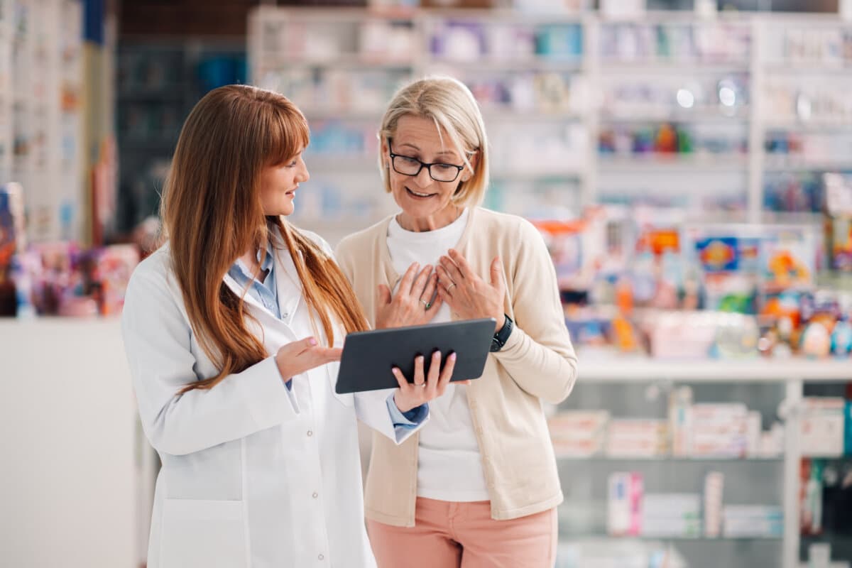 Smiling pharmacist is showing information on a digital tablet to a surprised senior customer while standing in a pharmacy